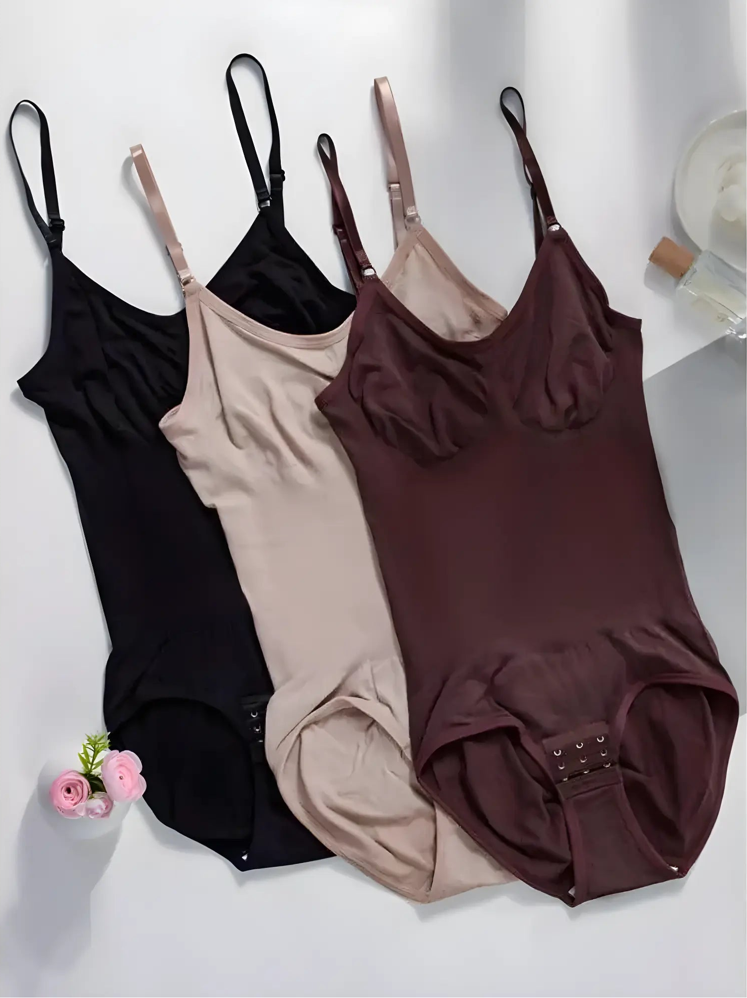 Vertical arrangement of Velmora bodysuits in neutral tones on a clean white surface, accompanied by minimalist decor to represent luxury shapewear for the modern woman.